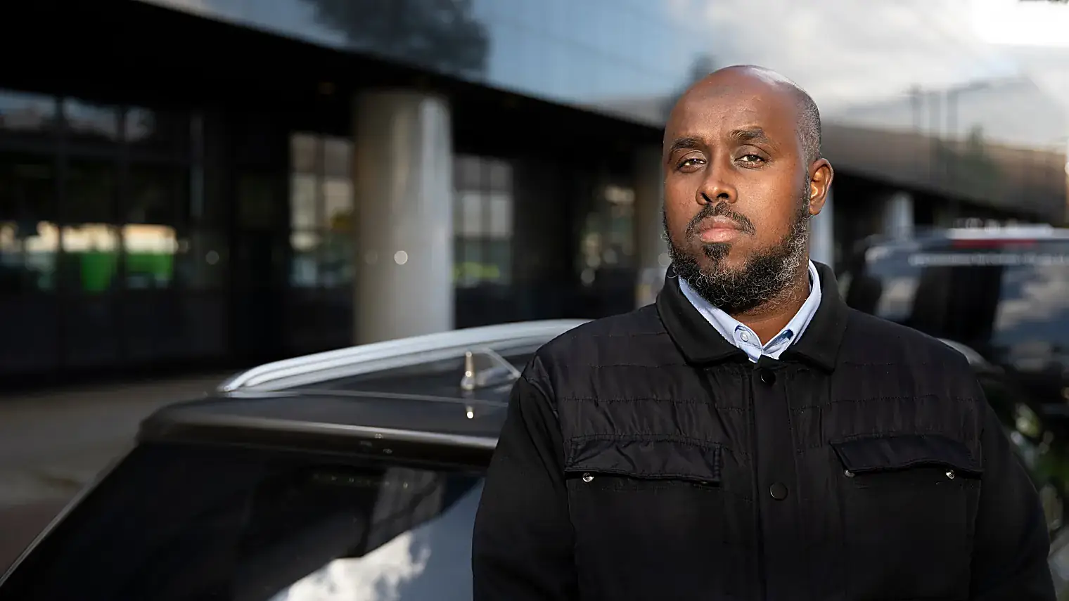 Uber-chauffeur Mohammed Shirwa in een zwarte jas naast zijn donkere auto voor een glazen kantoorgebouw in Rotterdam