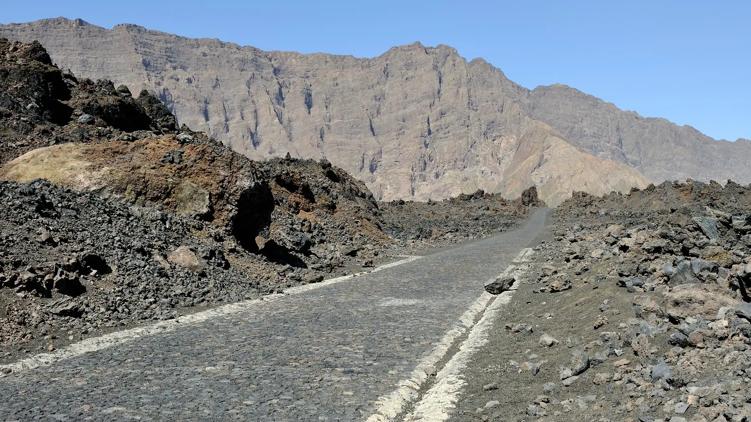 Onverharde weg door een zwart lavaveld in de krater van de vulkaan Pico do Fogo, met kale bergwanden op de achtergrond