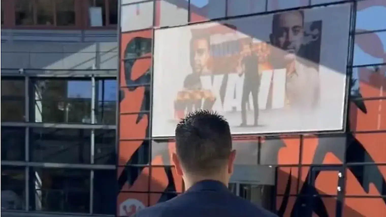 Xavi Hernandez van achteren gezien terwijl hij naar het gebouw van de KNVB Campus loopt, met op de gevel een groot scherm met zijn eigen naam en foto