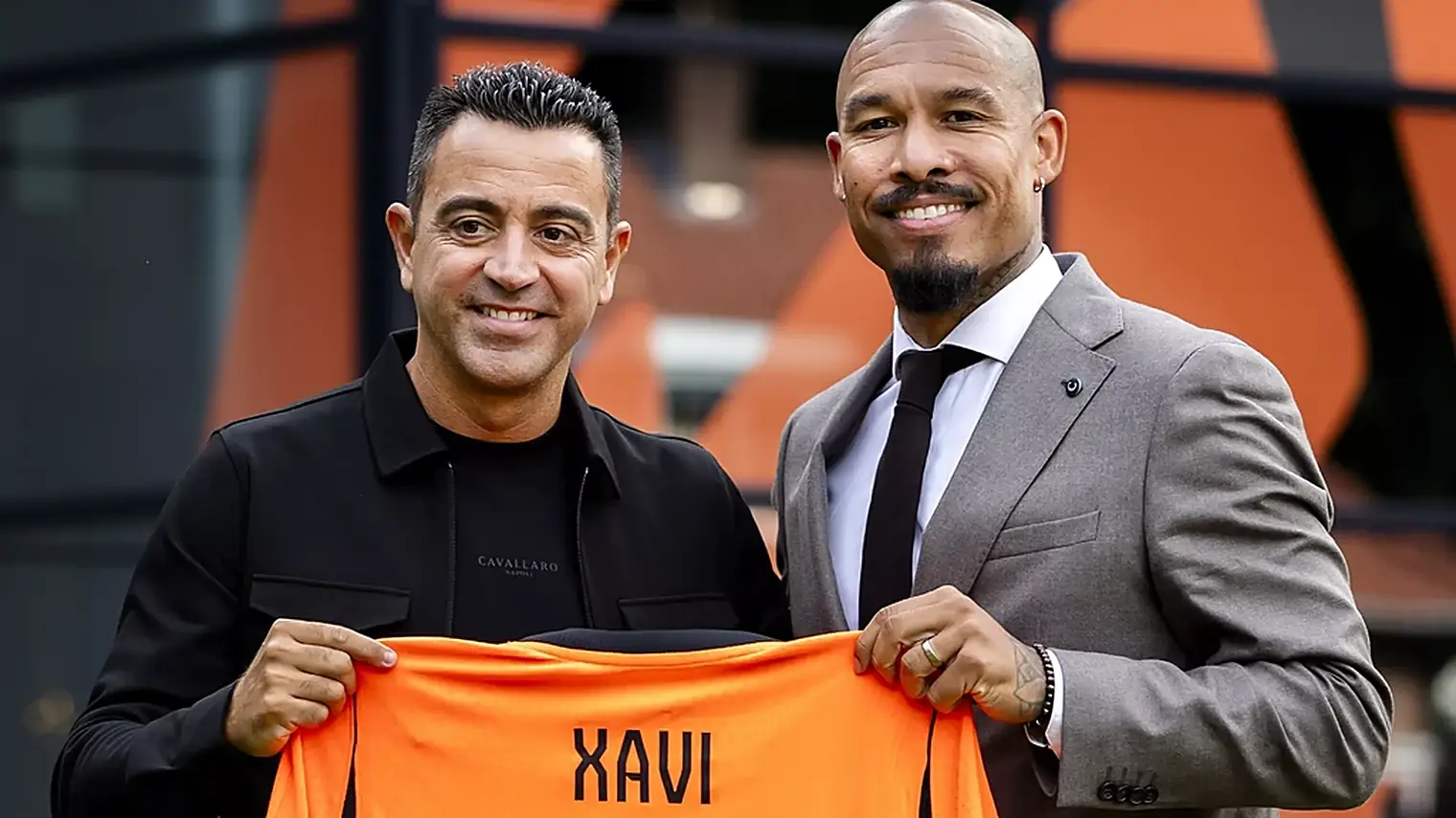 Xavi Hernandez en Nigel de Jong houden lachend een oranje shirt omhoog met de naam Xavi op de achterkant, buiten op de KNVB Campus