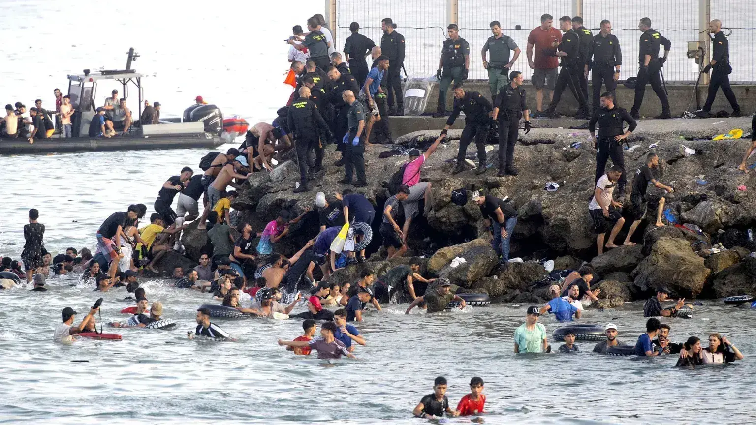 Honderden mensen zwemmen en waden rond de rotsen bij de pier van Ceuta, terwijl agenten van de Guardia Civil vanaf de kade en een boot toekijken