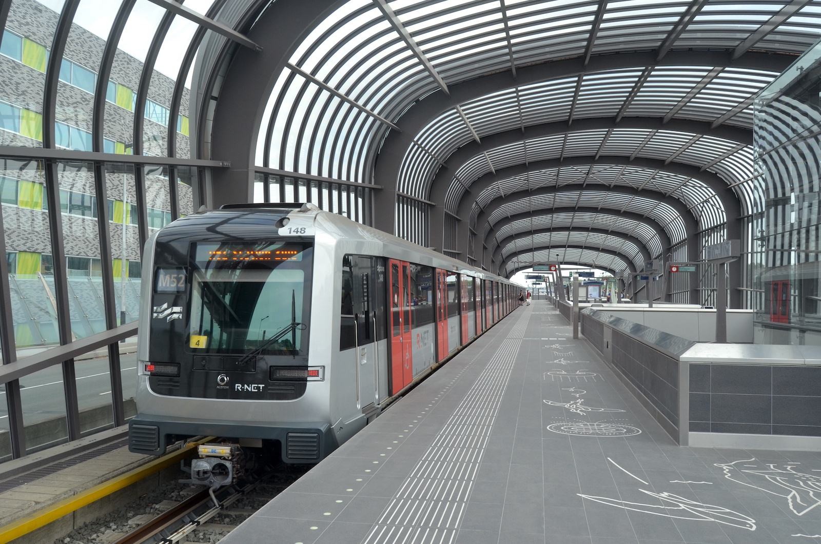 Een metro van lijn M52 staat bij het perron van een station met een gebogen glazen overkapping