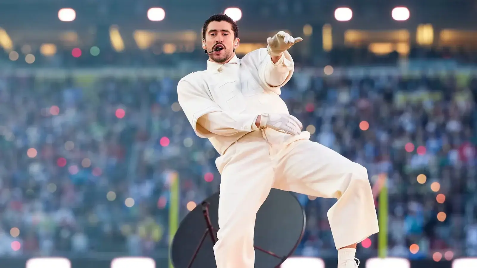 Bad Bunny in een wit trainingspak danst op het podium tijdens zijn Super Bowl-halftimeshow, met een vol, wazig stadion op de achtergrond