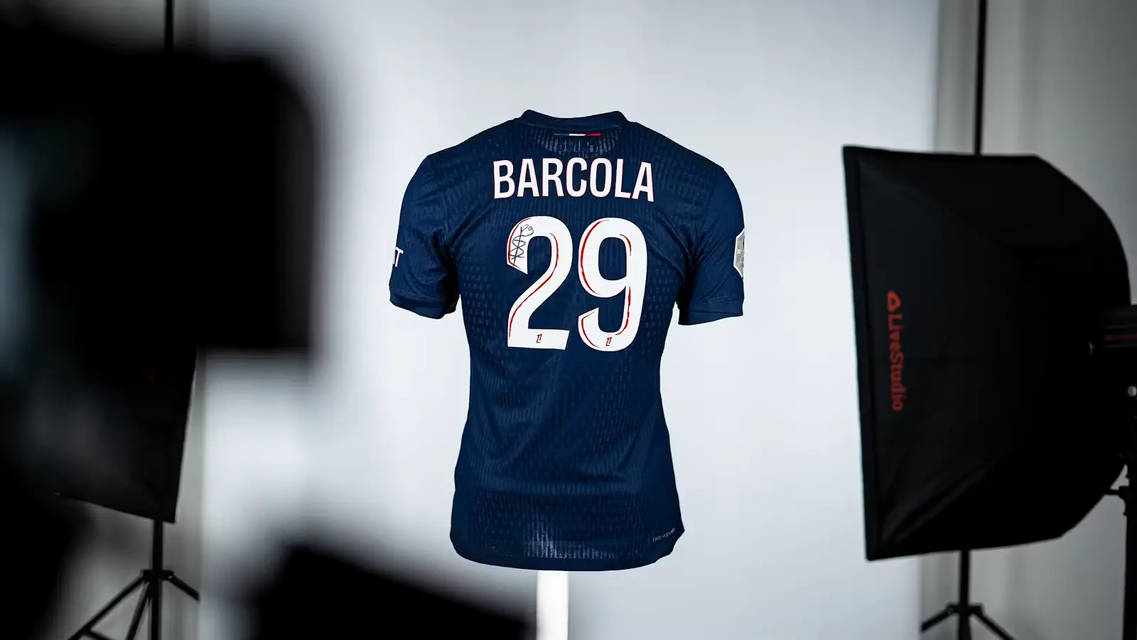Gesigneerd donkerblauw PSG-shirt met de naam Barcola en rugnummer 29, opgehangen in een fotostudio met softboxen