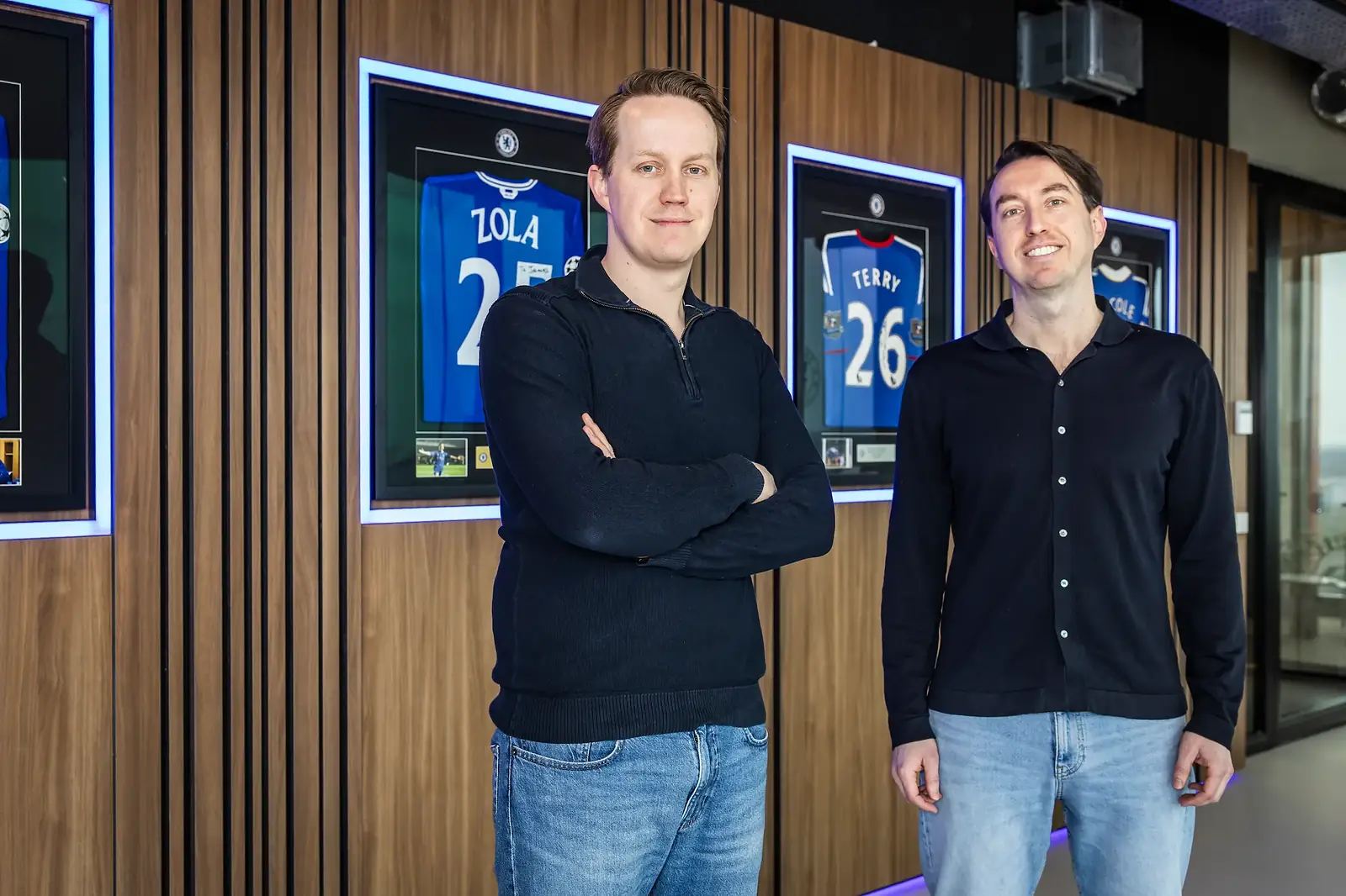 Bob en Tijmen Zonderwijk van MatchWornShirt staan voor een houten wand met ingelijste Chelsea-shirts van Zola en Terry