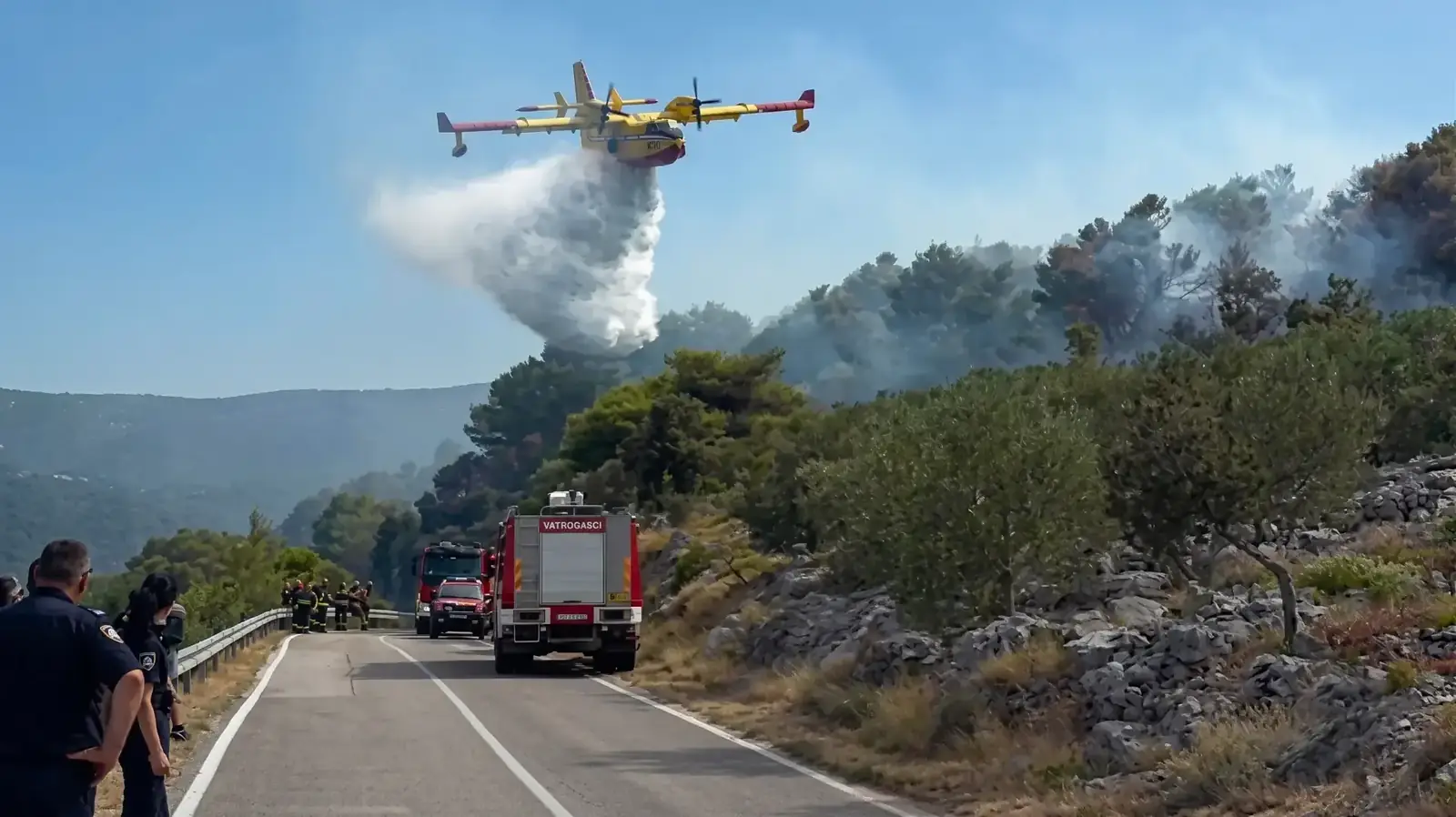 Een Canadair-blusvliegtuig laat water vallen boven een brand langs een weg, met Kroatische brandweerwagens en agenten op de voorgrond