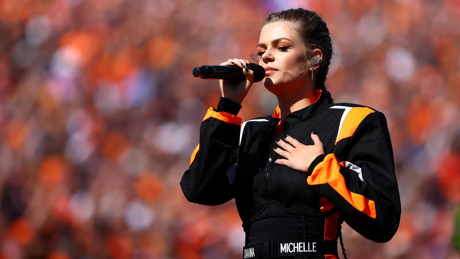 Davina Michelle zingt met een hand op haar hart in een zwart-oranje racepak het volkslied, voor een oranje gekleurde tribune