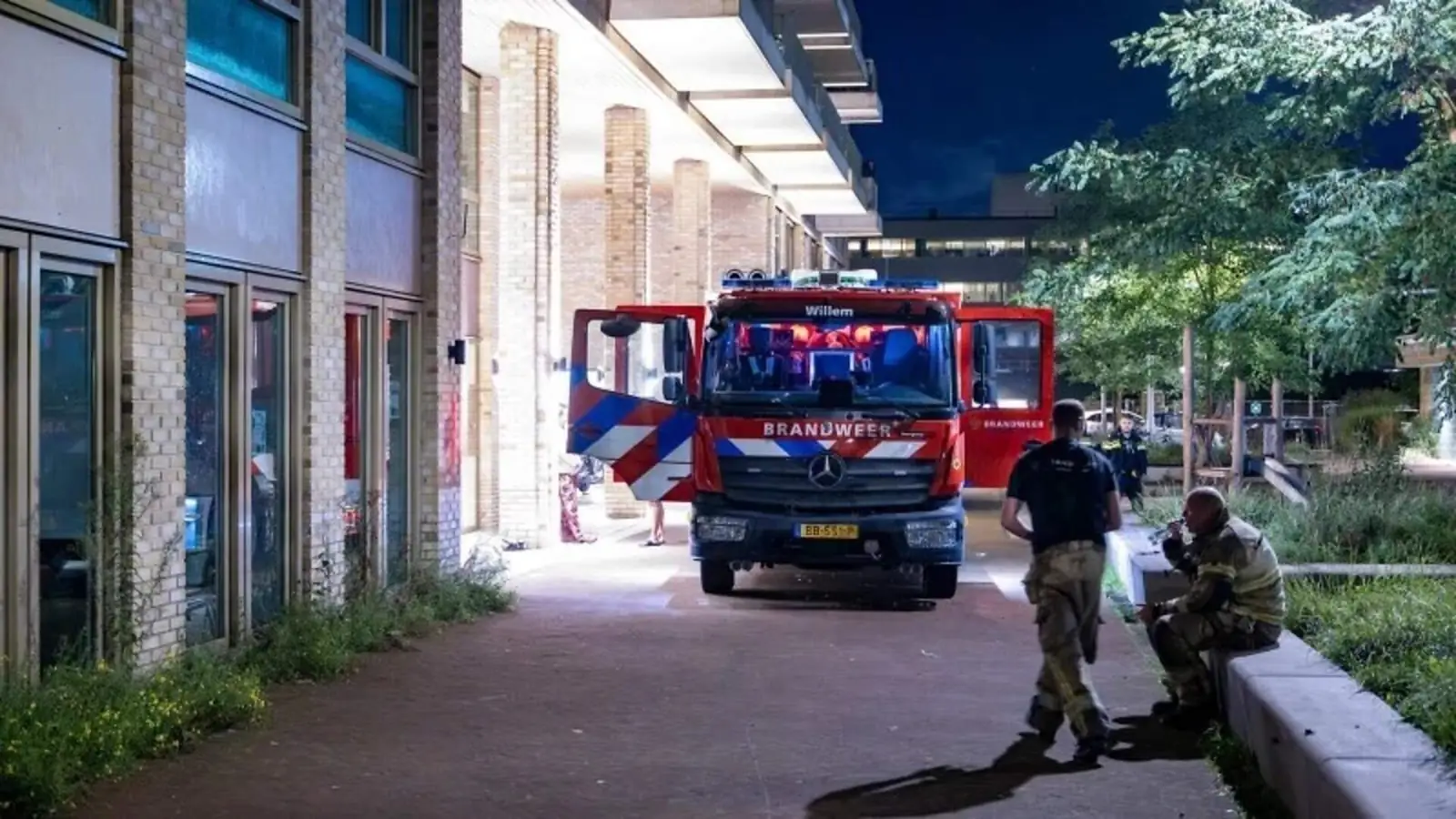 Een brandweerwagen van de Amsterdamse brandweer staat s nachts voor een gebouw, met twee brandweermannen ervoor