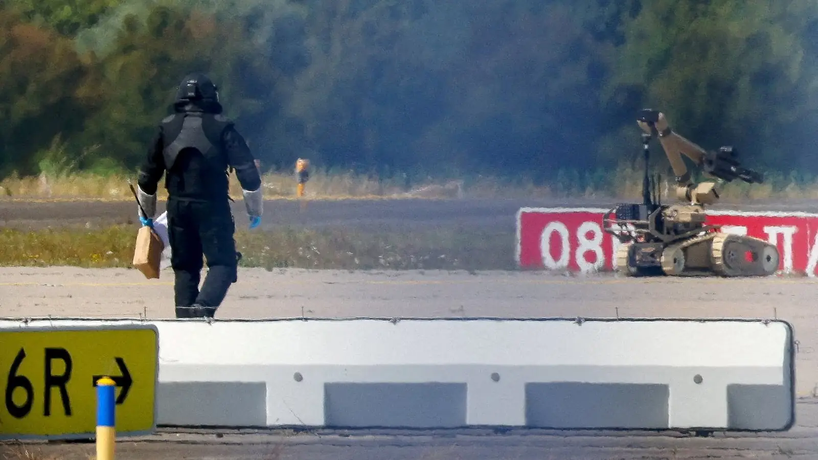 Een lid van de explosievenopruimingsdienst in beschermend pak loopt met een pakket over de startbaan, met een rupsrobot op de achtergrond