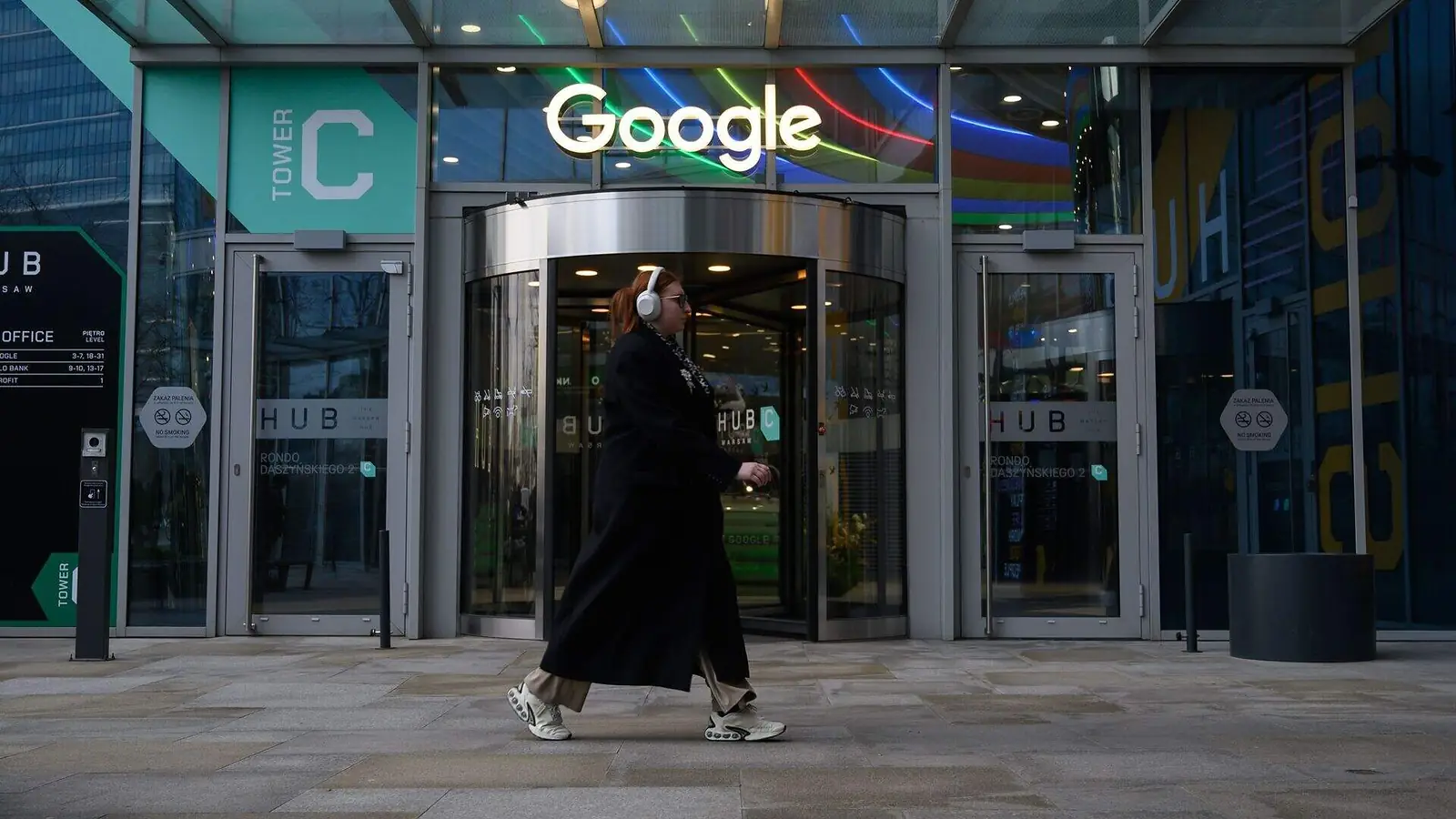 Een vrouw met koptelefoon loopt langs de ingang van een Google-kantoor, met het lichtgevende Google-logo boven de deur