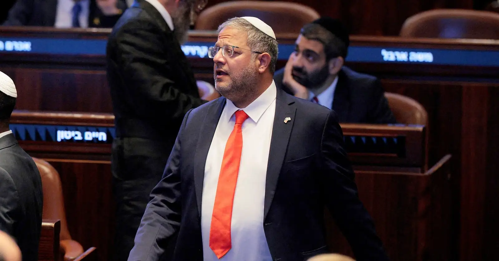 Itamar Ben-Gvir met keppel en rode stropdas loopt door de plenaire zaal van de Knesset, met parlementsleden op de achtergrond