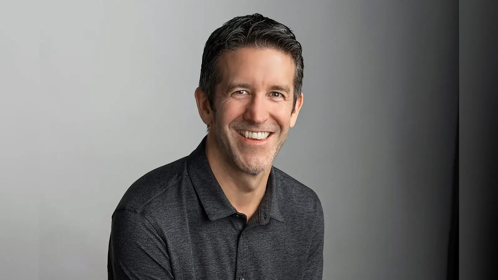 Portret van John Ternus, de nieuwe CEO van Apple, lachend tegen een grijze achtergrond