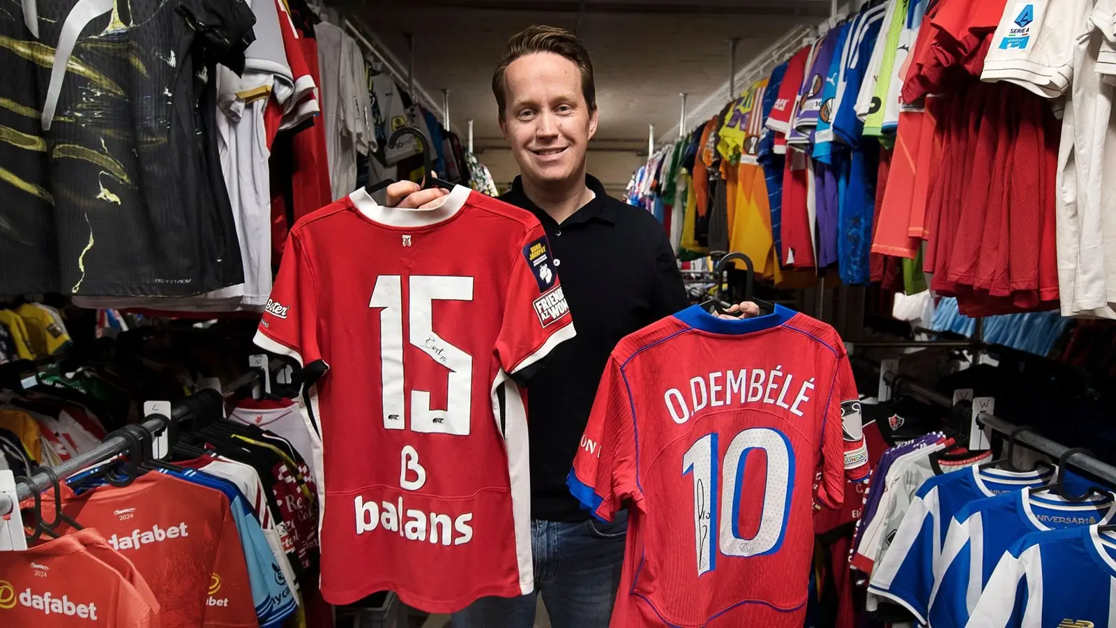 Medeoprichter Bob Zonderwijk van MatchWornShirt houdt twee gesigneerde voetbalshirts op in een magazijn met lange rekken vol shirts