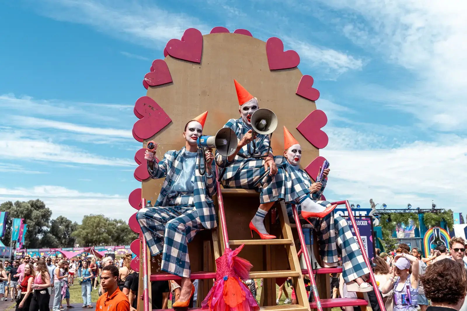 Drie performers met witte clownsmake-up en oranje puntmutsen in blauw geblokte pakken op een trap voor een decor met roze harten, met de drukte van Milkshake Festival op de achtergrond