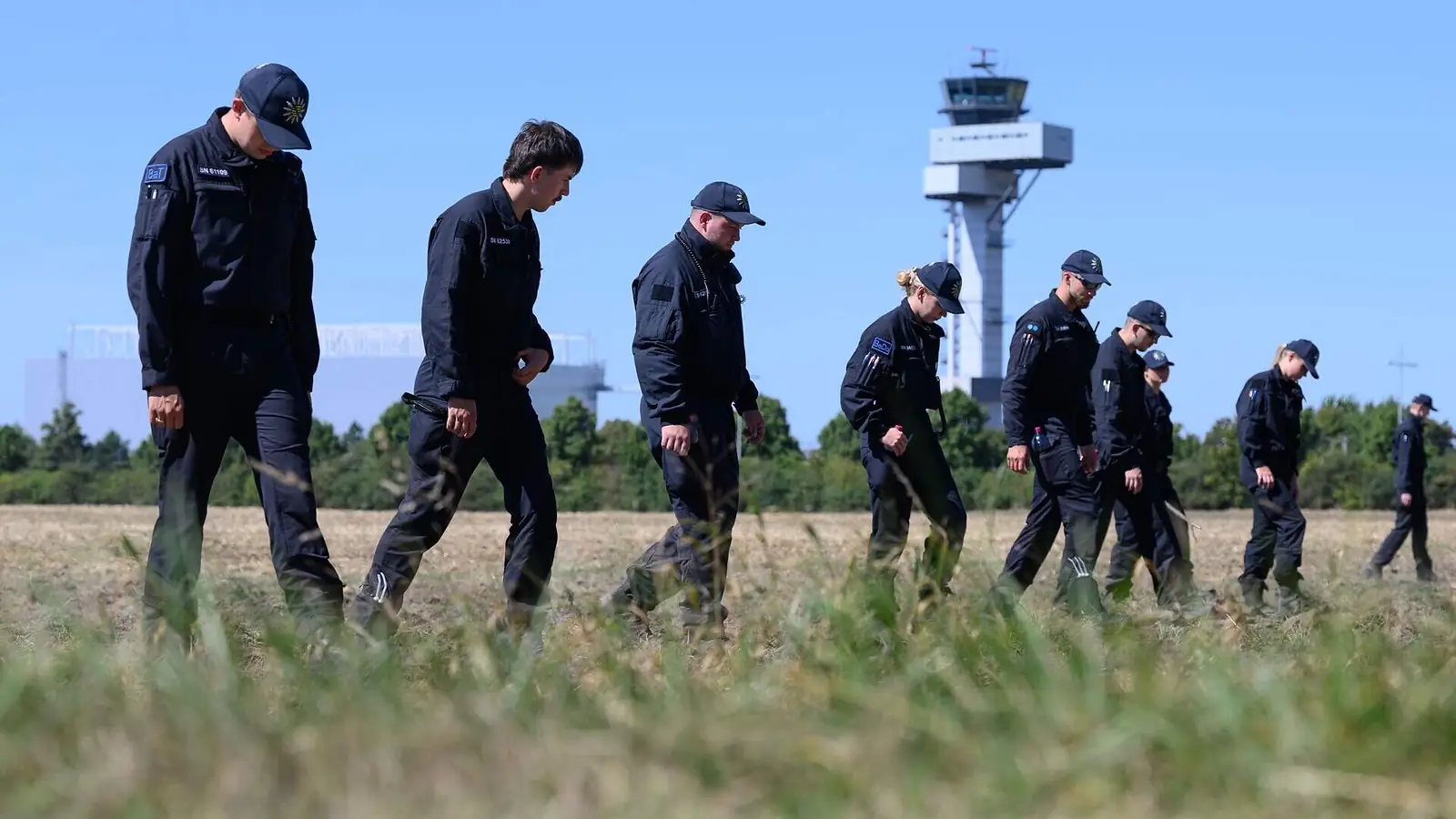 Een rij Duitse agenten in donkere uniformen loopt zoekend door een veld, met op de achtergrond de verkeerstoren van de luchthaven van Leipzig