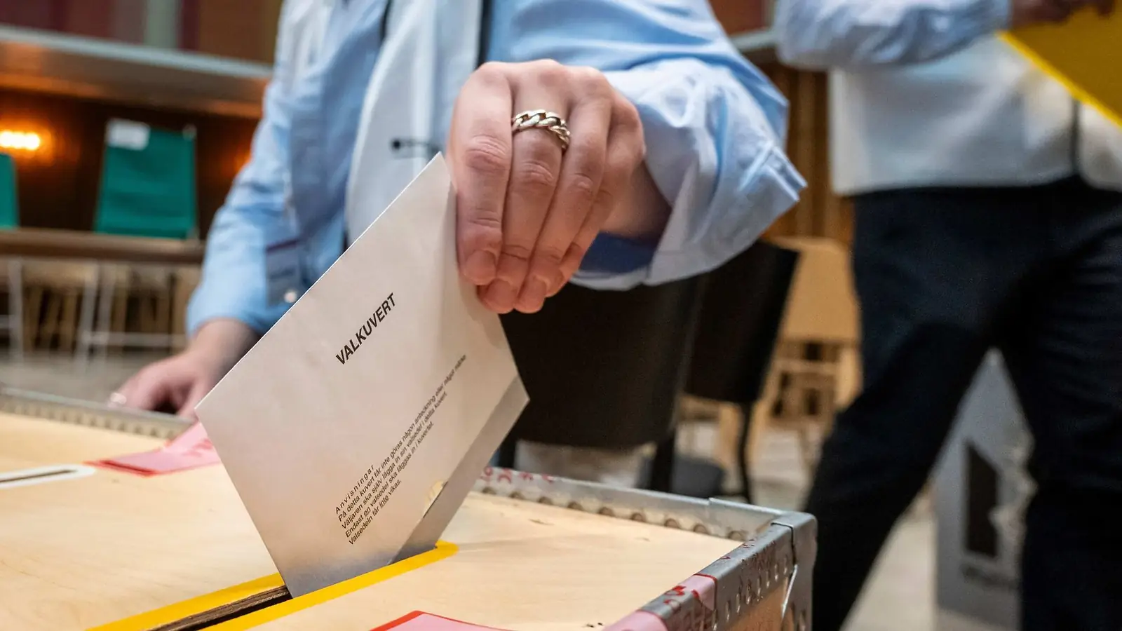 Een hand met ring stopt een envelop met opschrift "Valkuvert" in een houten stembus in een Zweeds stembureau