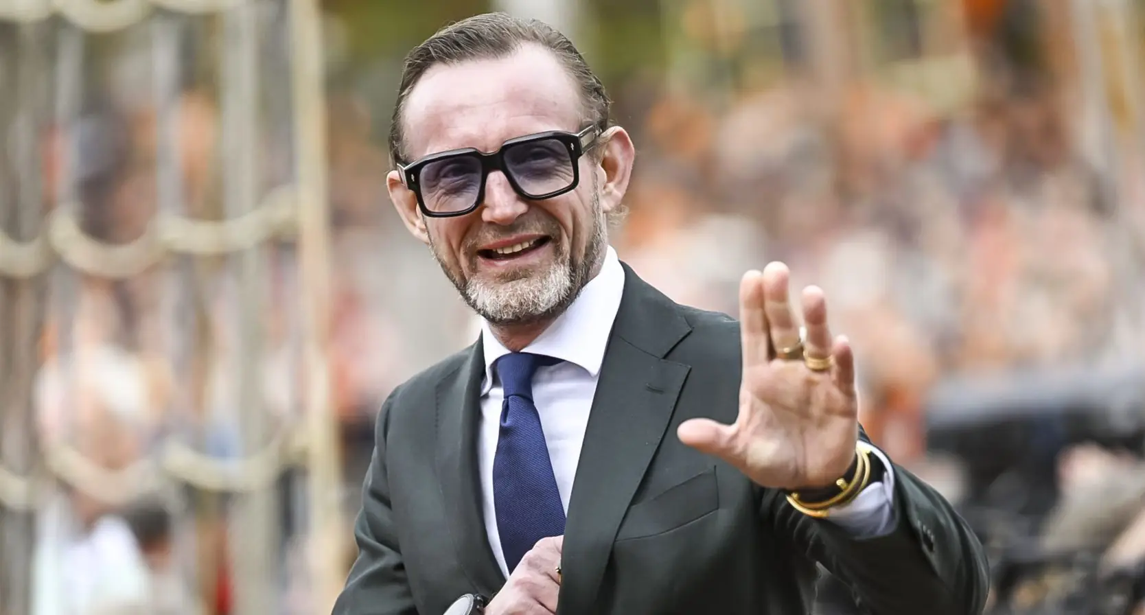 Prins Bernhard van Oranje in donker pak met zonnebril, zwaaiend naar publiek langs een afzetting