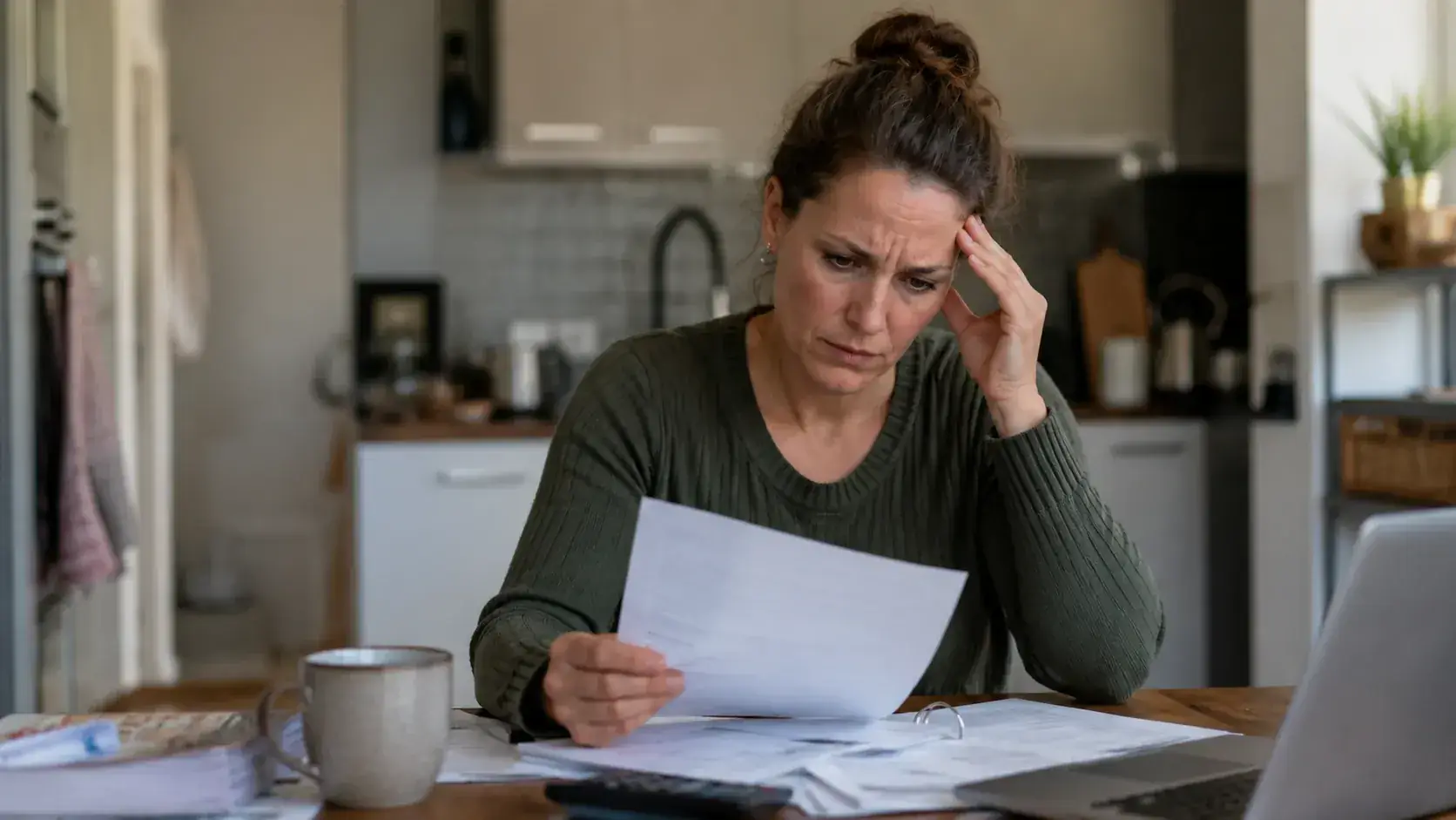Vrouw zit aan een keukentafel met papieren, een rekenmachine en een laptop voor zich, met haar hand tegen haar hoofd