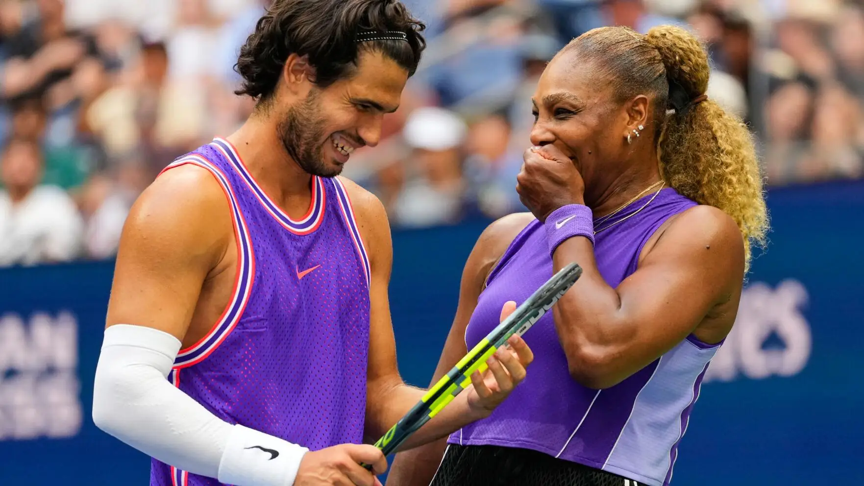 Carlos Alcaraz en Serena Williams lachen naar elkaar in paars tenue op de baan, met een volle tribune achter hen