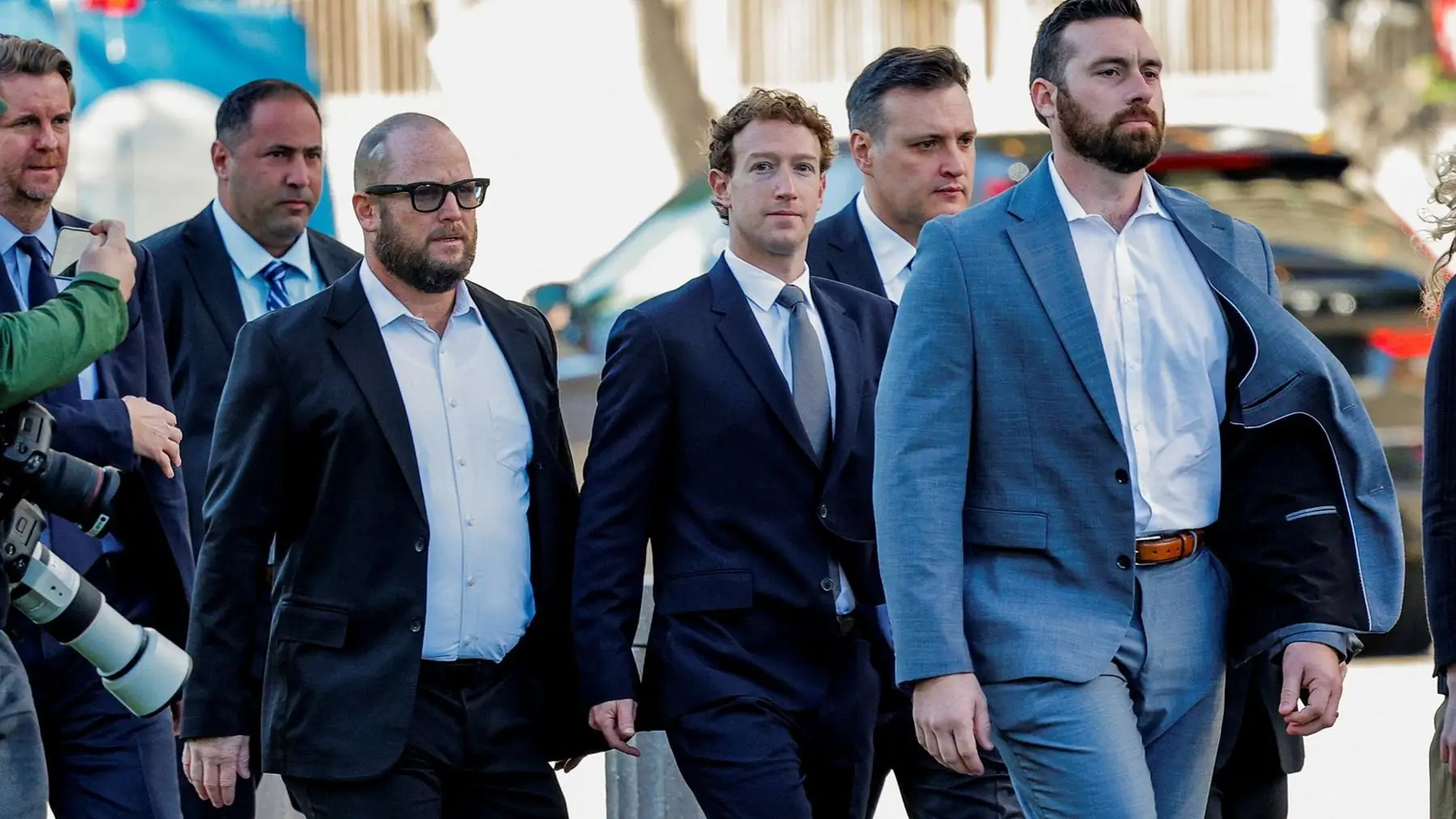 Mark Zuckerberg loopt in donker pak tussen beveiligers en medewerkers over straat, links op de voorgrond een fotograaf met telelens