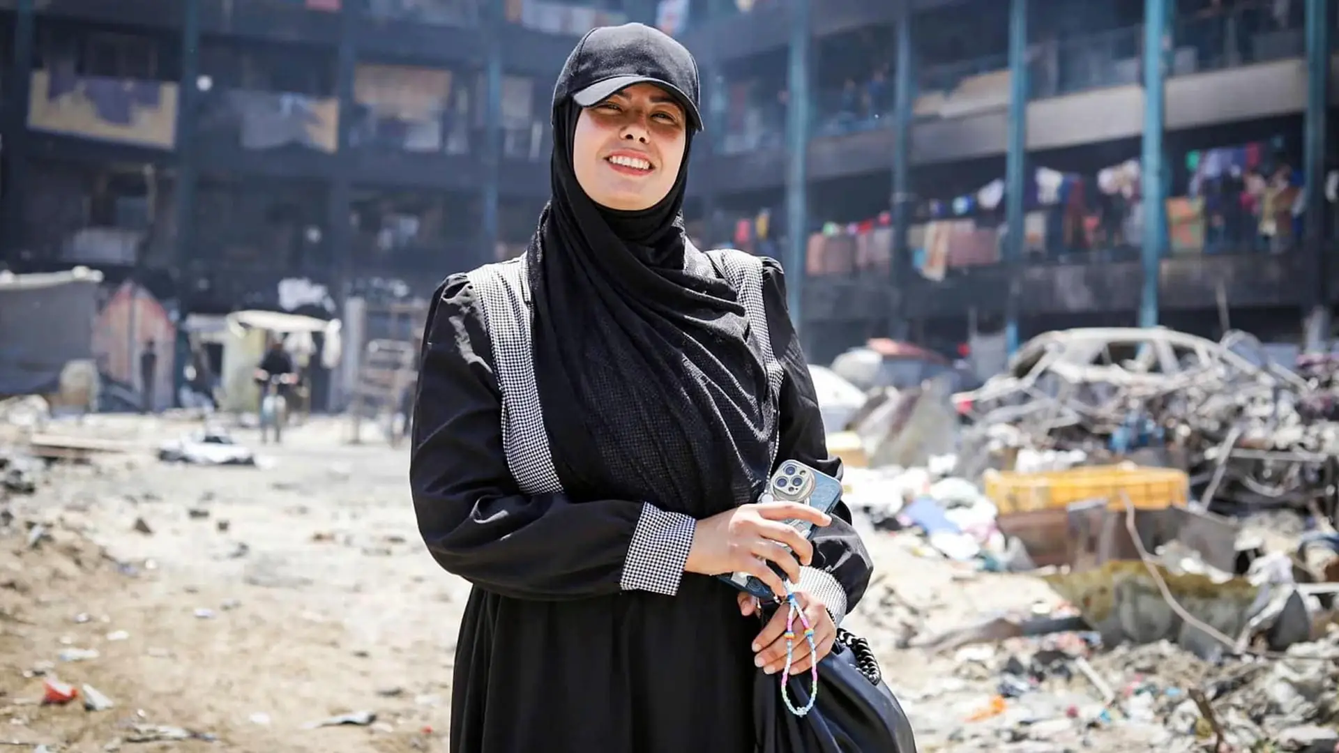 Fatma Hassona staat lachend met haar telefoon in de hand op een zandpad tussen puin en een uitgebrand gebouw in Gaza-Stad