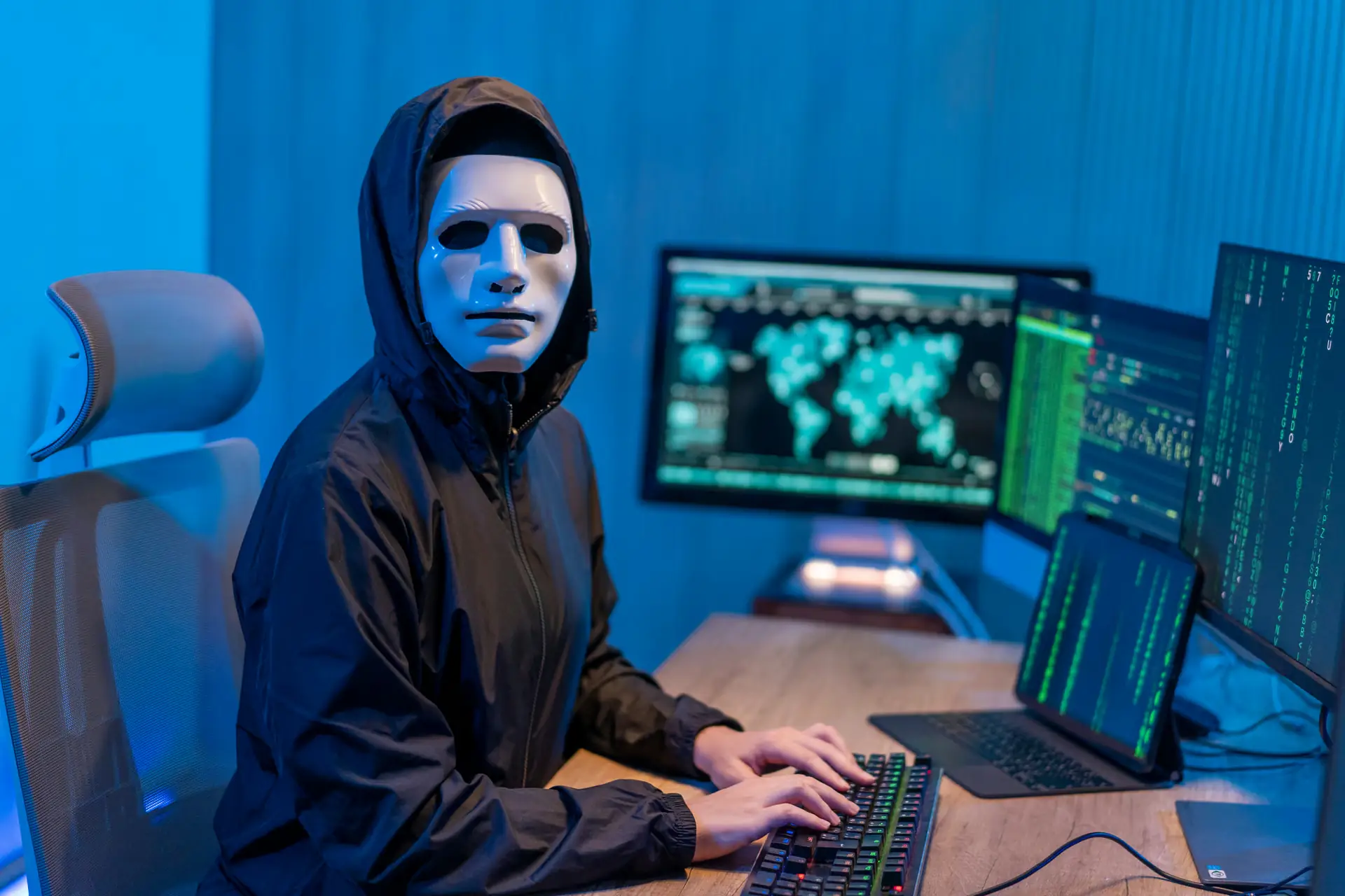 Iemand met een wit masker en een zwarte capuchon typt op een toetsenbord voor beeldschermen met een wereldkaart en groene datacode