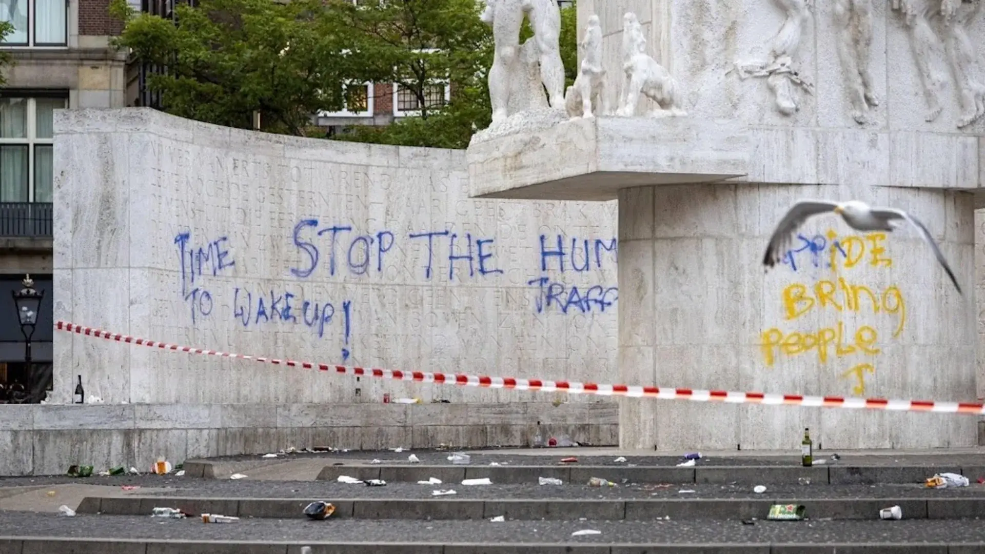 Blauwe graffiti met de teksten Time to wake up en Stop the human traffic op de muur van het Nationaal Monument op de Dam, rechts in geel de tekst Pride bring people, met rood-wit afzetlint ervoor