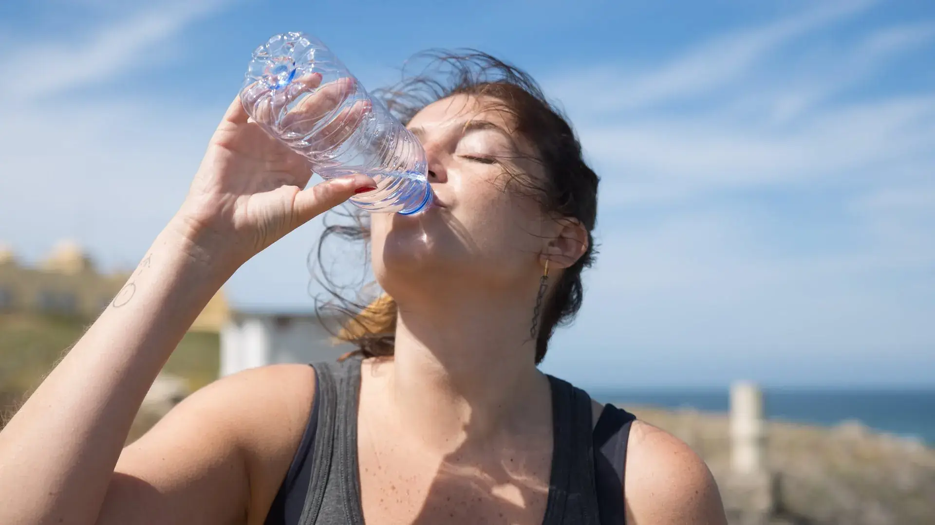 Een vrouw drinkt water uit een plastic fles in de volle zon, met een strakblauwe lucht en de zee op de achtergrond
