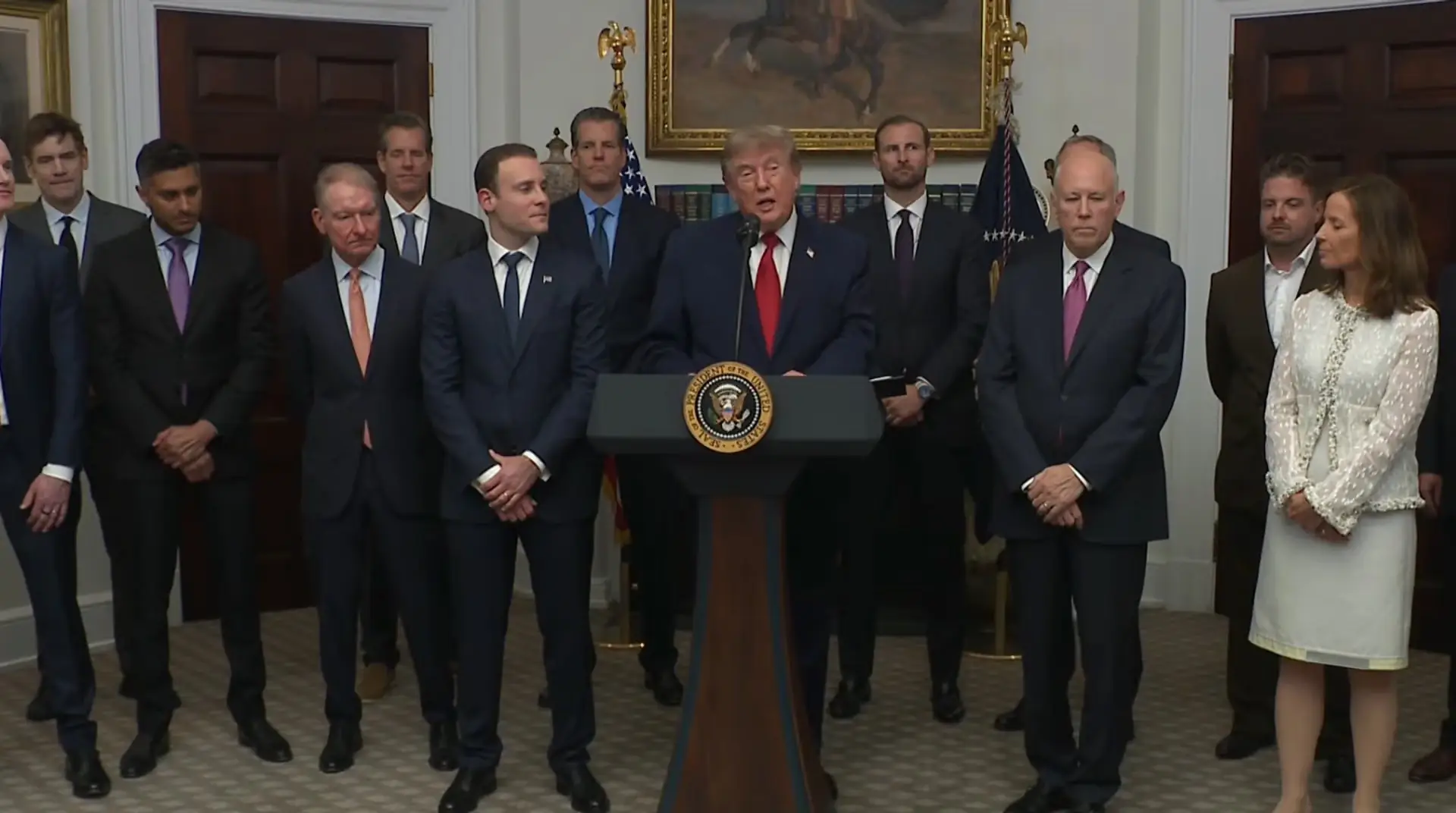 President Trump spreekt achter een katheder met het presidentieel zegel in het Witte Huis, omringd door een groep mannen en vrouwen in pak