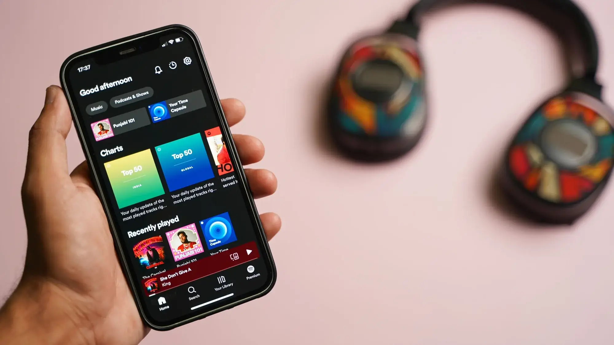 Hand houdt een telefoon met de Spotify-app open op de hitlijsten, met een koptelefoon onscherp op een roze achtergrond