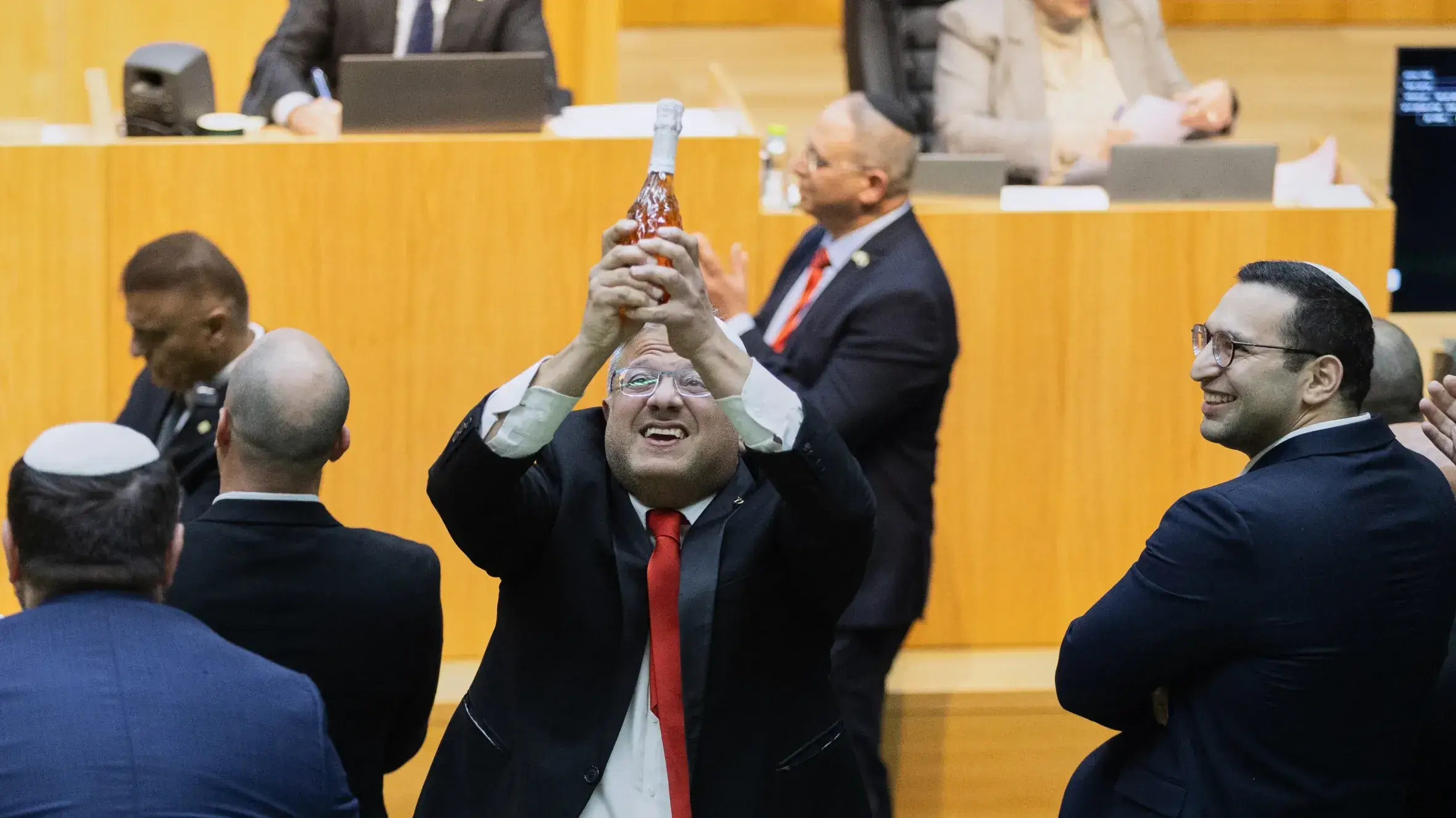 Ben-Gvir houdt lachend een fles champagne omhoog in de plenaire zaal van de Knesset, omringd door applaudisserende parlementsleden