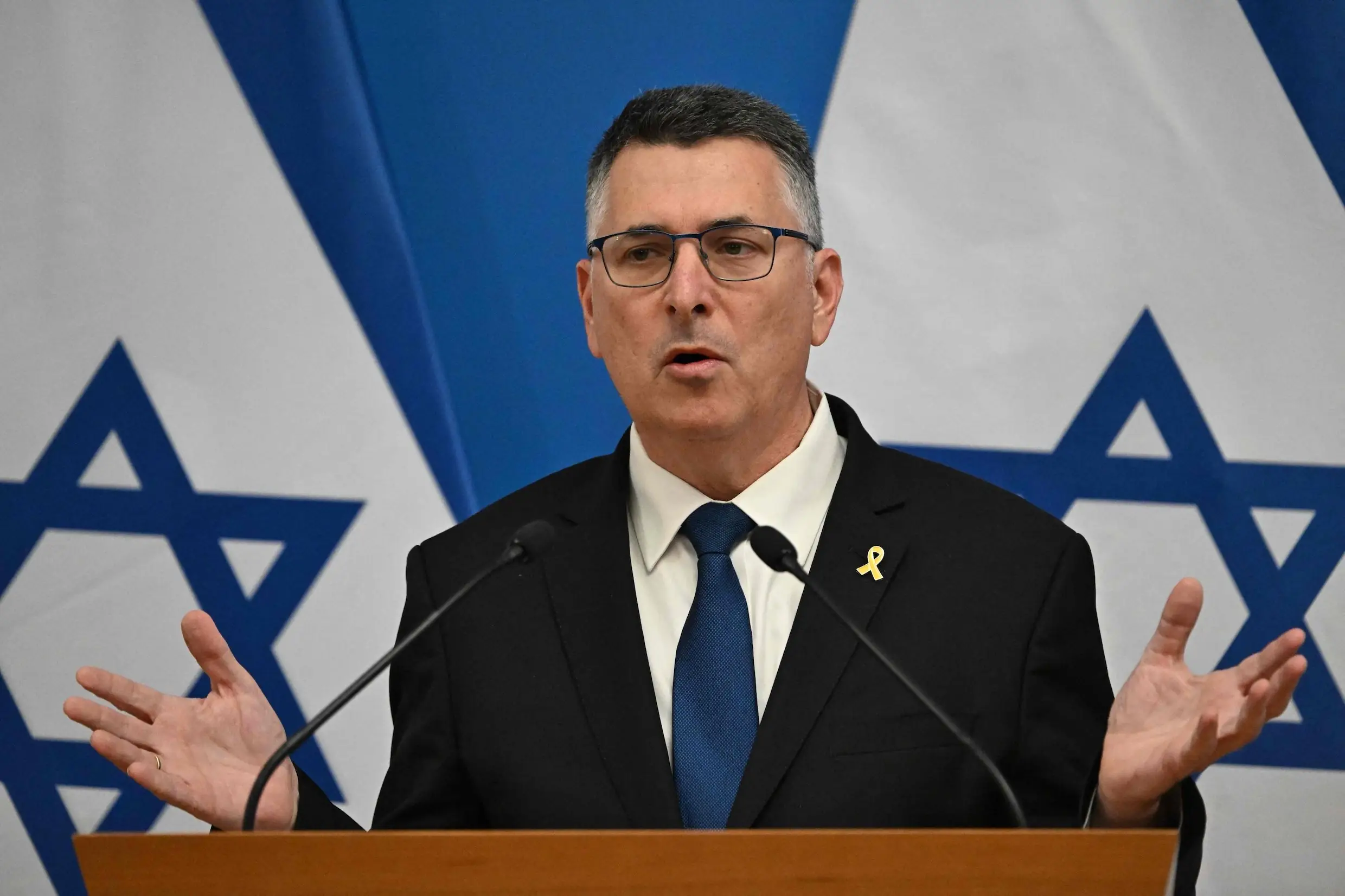De Israelische minister van Buitenlandse Zaken Gideon Saar spreekt met gespreide handen achter een katheder, voor twee Israelische vlaggen
