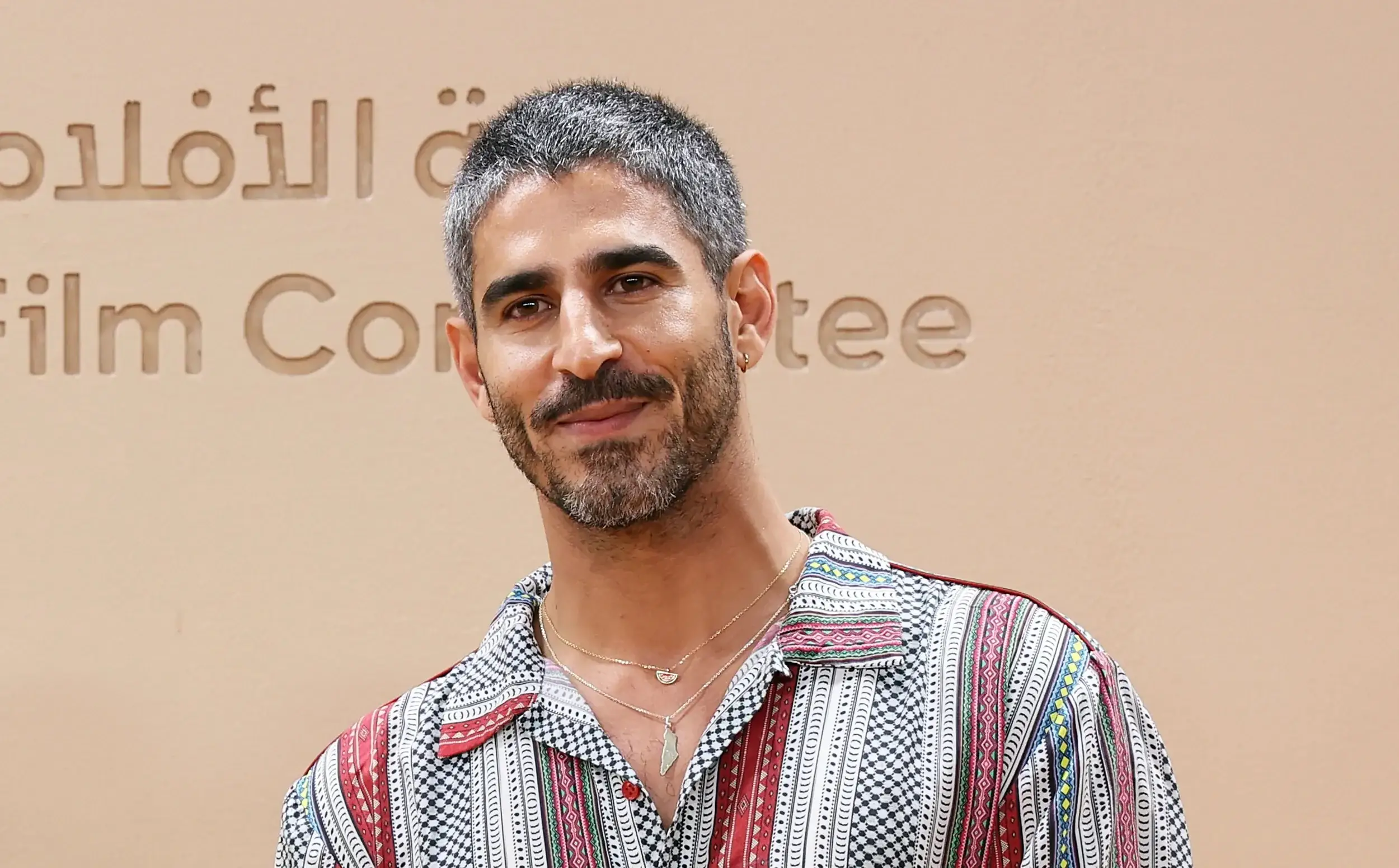 Ahmed Shihab-Eldin kijkt in de camera voor een zandkleurige wand met de tekst Film Committee, in een hemd met geometrisch patroon en een hanger in de vorm van Palestina