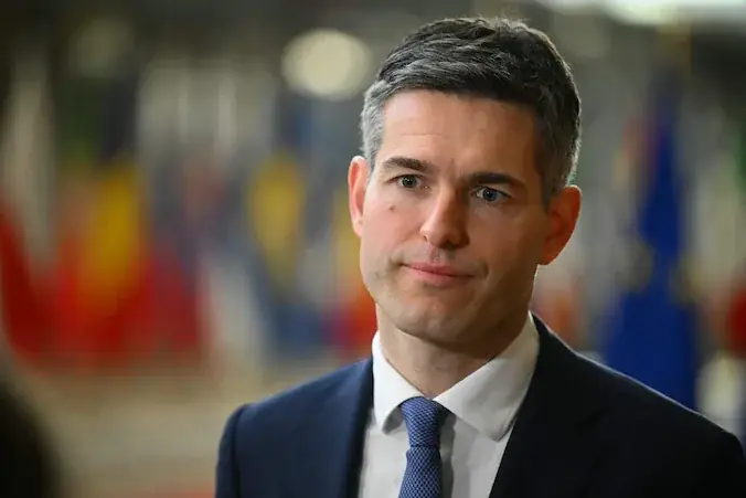 Minister Berendsen van Buitenlandse Zaken staat in een gang van het EU-gebouw in Brussel, met onscherpe vlaggen van de lidstaten achter hem