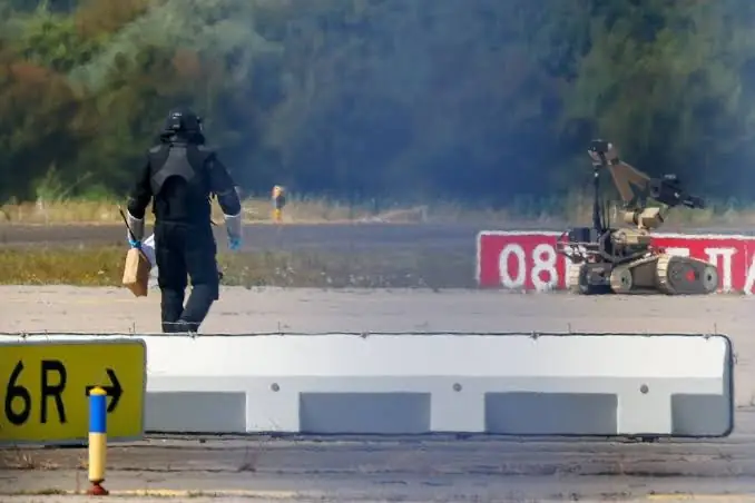 Een lid van de explosievenopruimingsdienst in beschermend pak loopt met een pakket over de startbaan, met een rupsrobot op de achtergrond