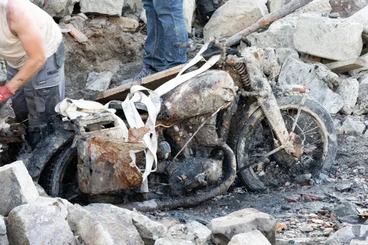 Zwaar verroeste motorfiets vol opgedroogd slib tussen de rotsblokken op de oever van de Donau, met spanbanden eromheen en twee mannen ernaast