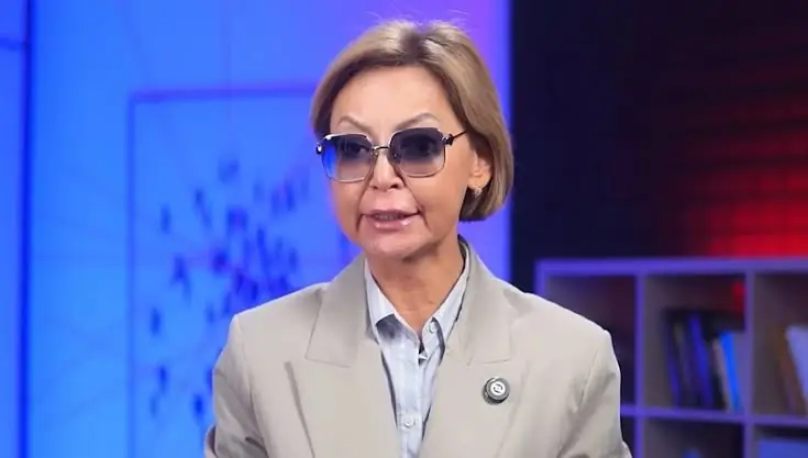 Gulnara Bazjkenova met getinte bril en kort blond haar in een televisiestudio, met blauw en rood licht op de achtergrond