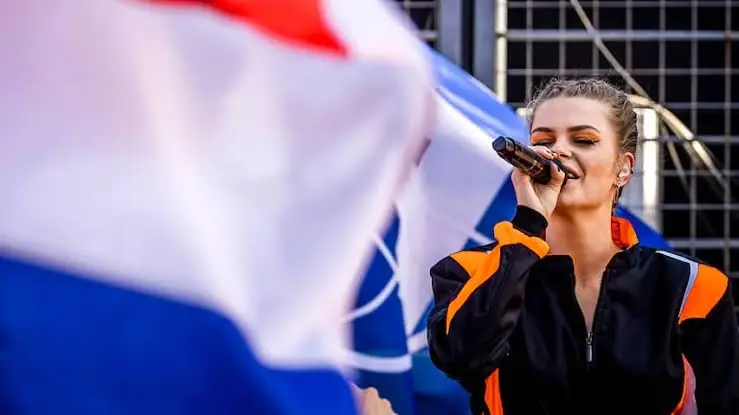 Davina Michelle zingt in een oranje-zwart racepak achter een wapperende Nederlandse vlag op het circuit