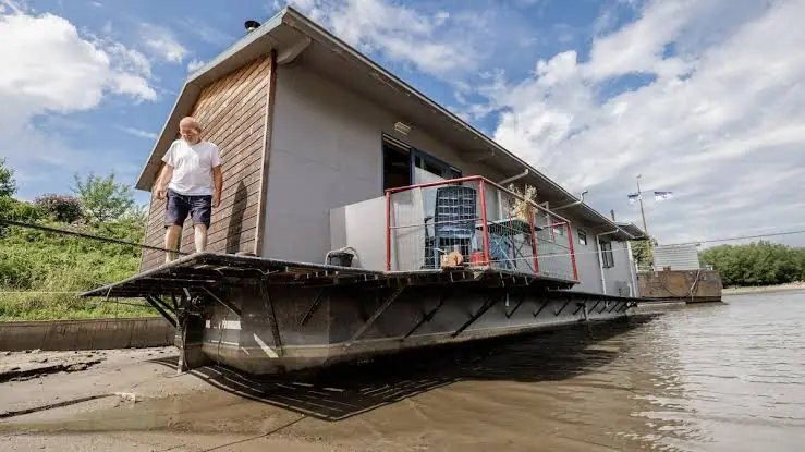 Man loopt over de loopplank van een woonark die op een drooggevallen, modderige oever langs de rivier ligt