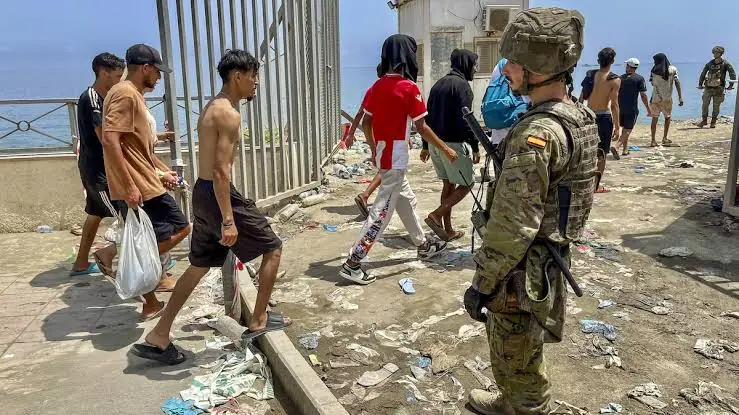 Een Spaanse militair met helm en geweer kijkt toe hoe een groep jonge mannen langs het grenshek van Ceuta terugloopt richting Marokko, over een strook vol afval
