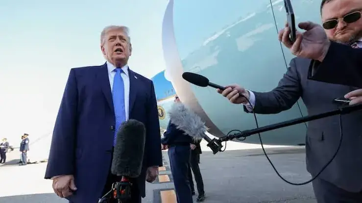 Donald Trump staat in donkerblauw pak voor een vliegtuig en beantwoordt vragen van journalisten die microfoons naar hem uitsteken