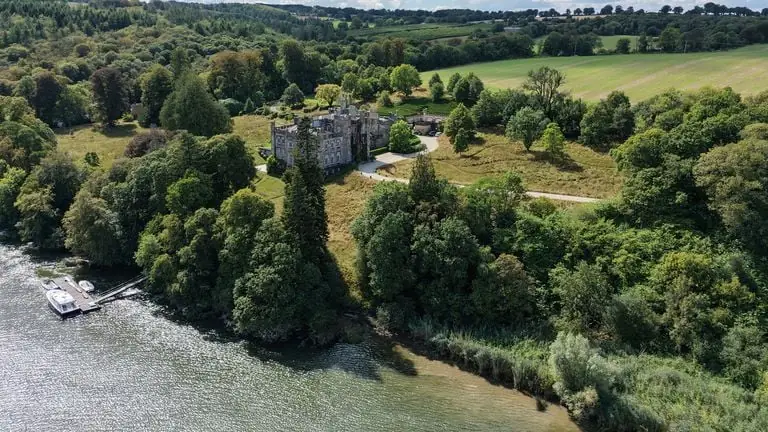 Luchtfoto van Strancally Castle tussen oude bomen aan de oever van de rivier Blackwater, met een aanlegsteiger en weilanden achter het huis