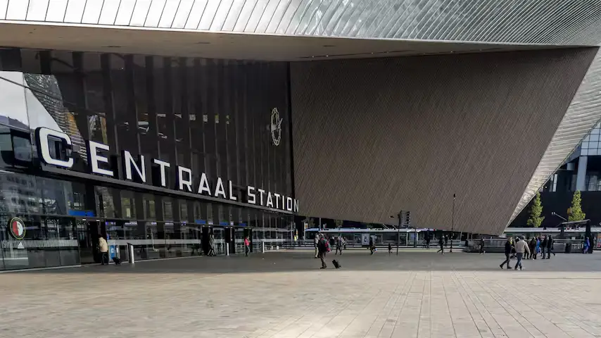 Voorgevel van Rotterdam Centraal met de letters Centraal Station onder de spitse luifel en reizigers op het stationsplein