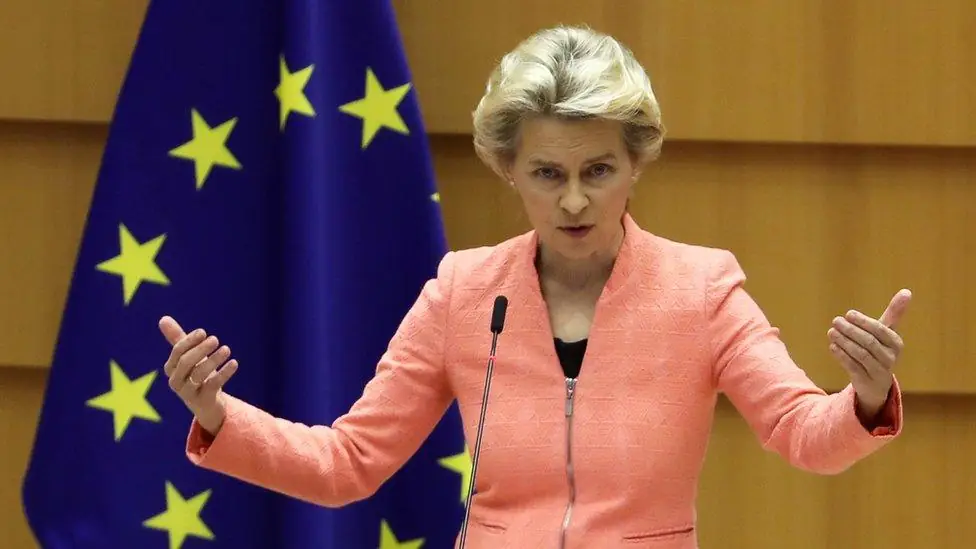 Commissievoorzitter Ursula von der Leyen spreekt achter een microfoon met beide handen omhoog, met de vlag van de Europese Unie achter haar