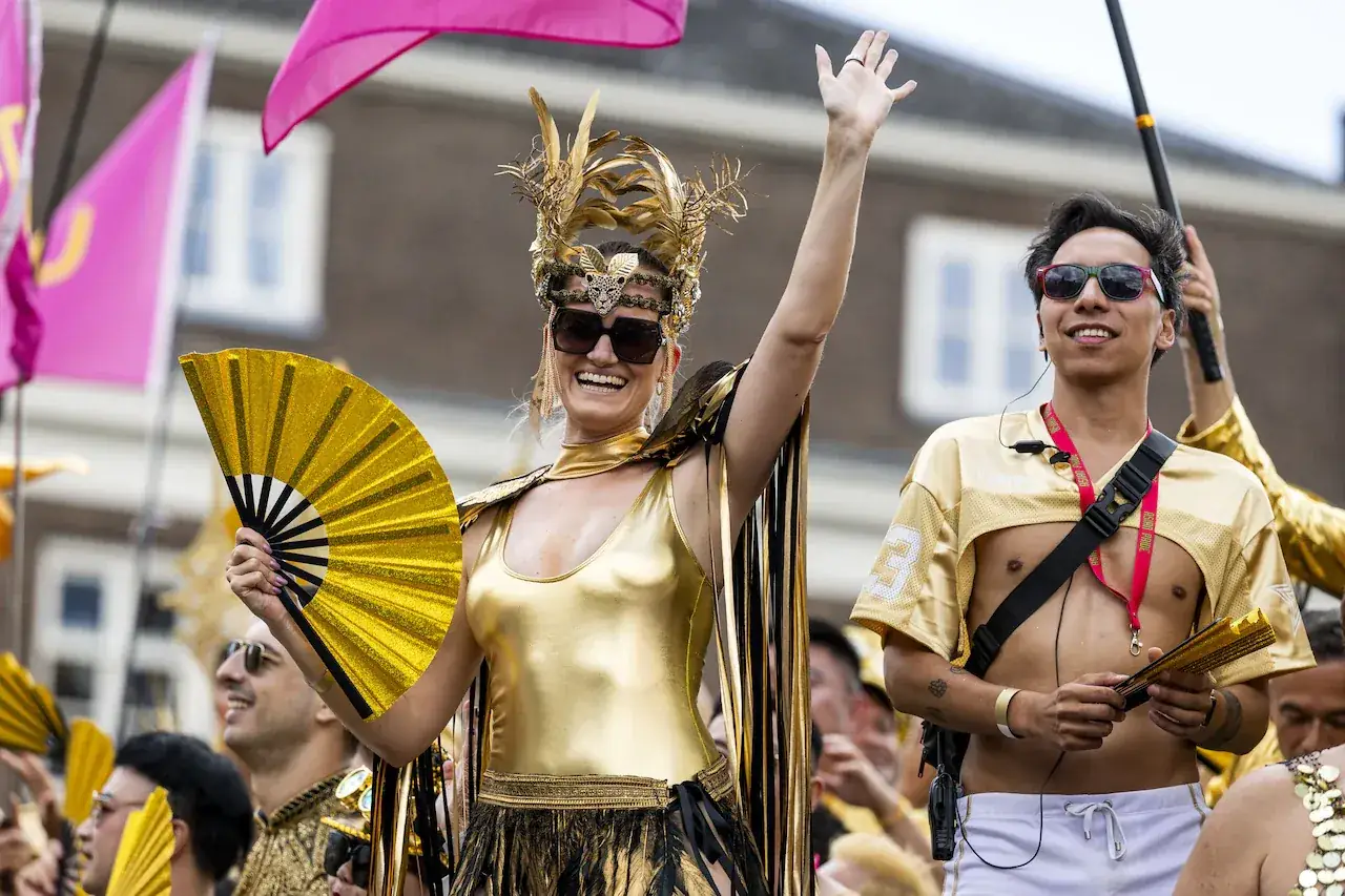 Twee feestvierders in gouden outfits met veren en waaiers zwaaien vanaf een boot in de Canal Parade