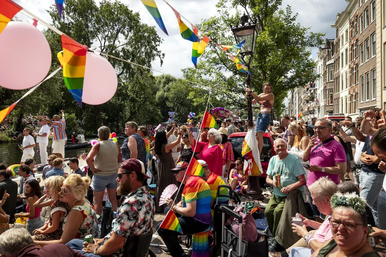 Publiek met regenboogvlaggen en ballonnen langs een Amsterdamse gracht kijkt naar de voorbijvarende boten
