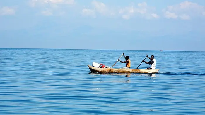 Twee mannen peddelen in een houten kano over het spiegelgladde Malawimeer, met in de verte de bergen aan de overkant